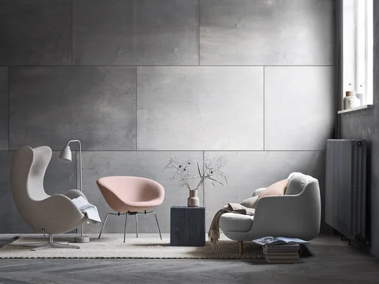 Le confortable canapé 2 places Lune de Fritz Hansen avec le fauteuil pot assorti dans des tons doux de gris et de rose. Une ambiance de salon moderne devant un mur gris.