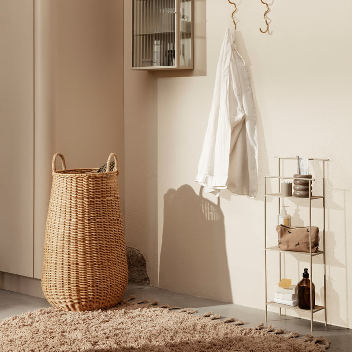 Le panier à linge en rotin Braided par ferm Vivre sur un tapis beige à côté d'une petite étagère avec des accessoires de salle de bains