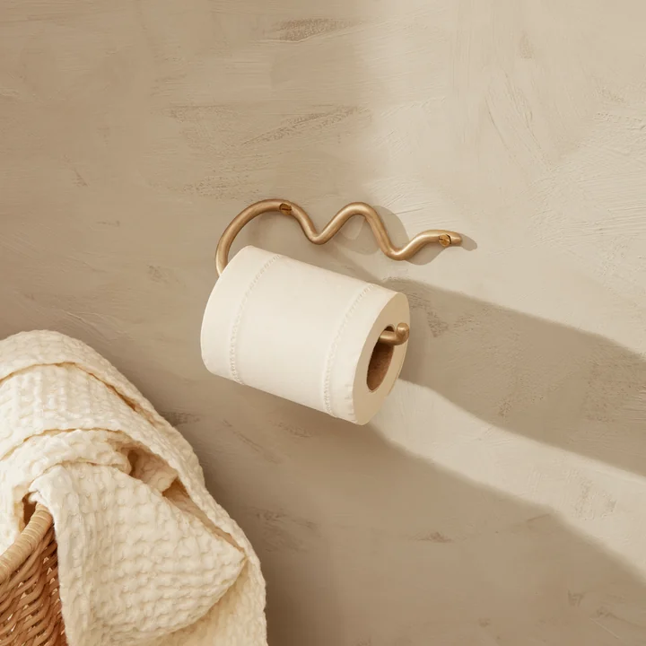 Le porte-papier hygiénique Curvature de Ferm Living sur un mur de pierre claire à côté d'un panier en rotin